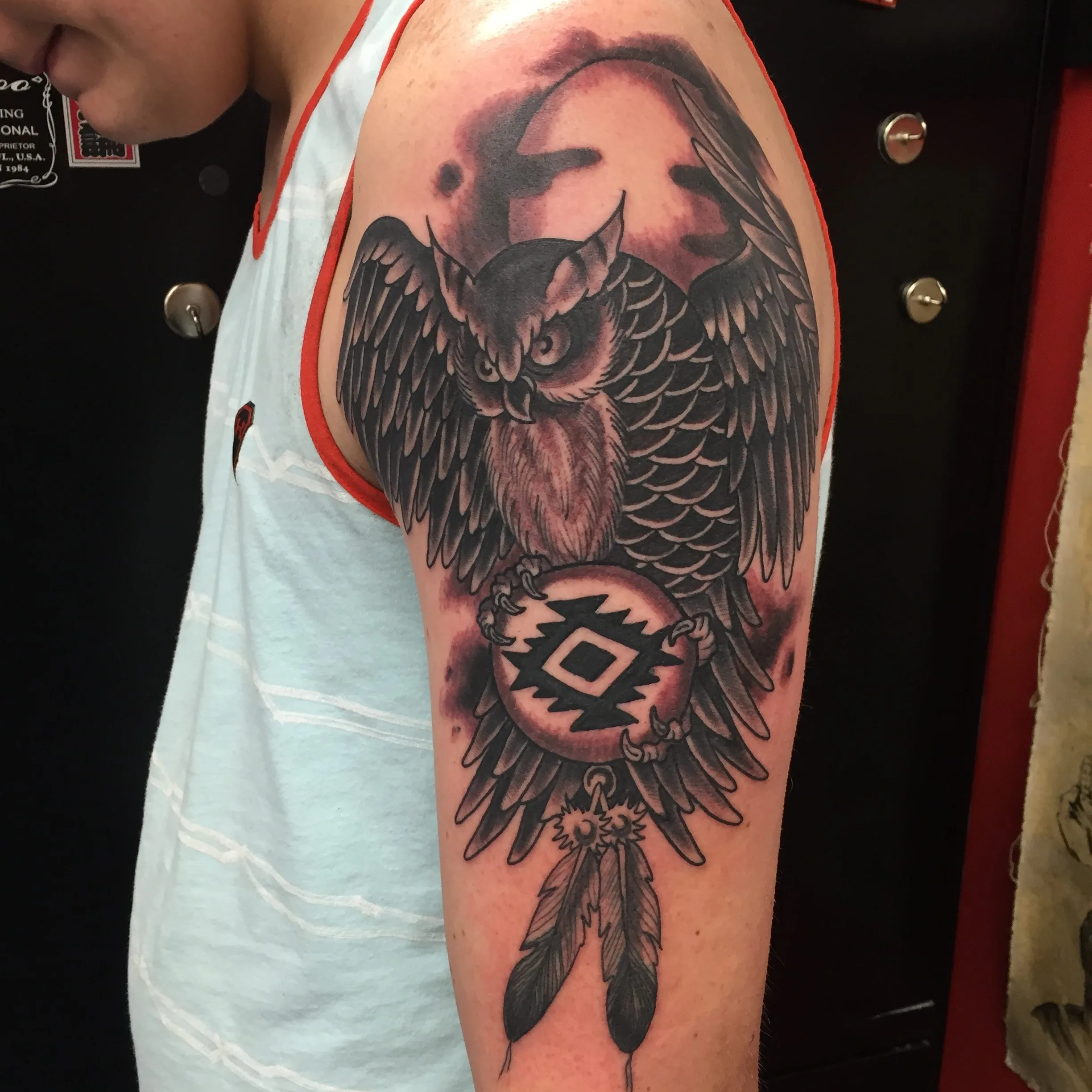Owl with spread wings above a round medallion design and hanging feathers, black and grey, upper arm