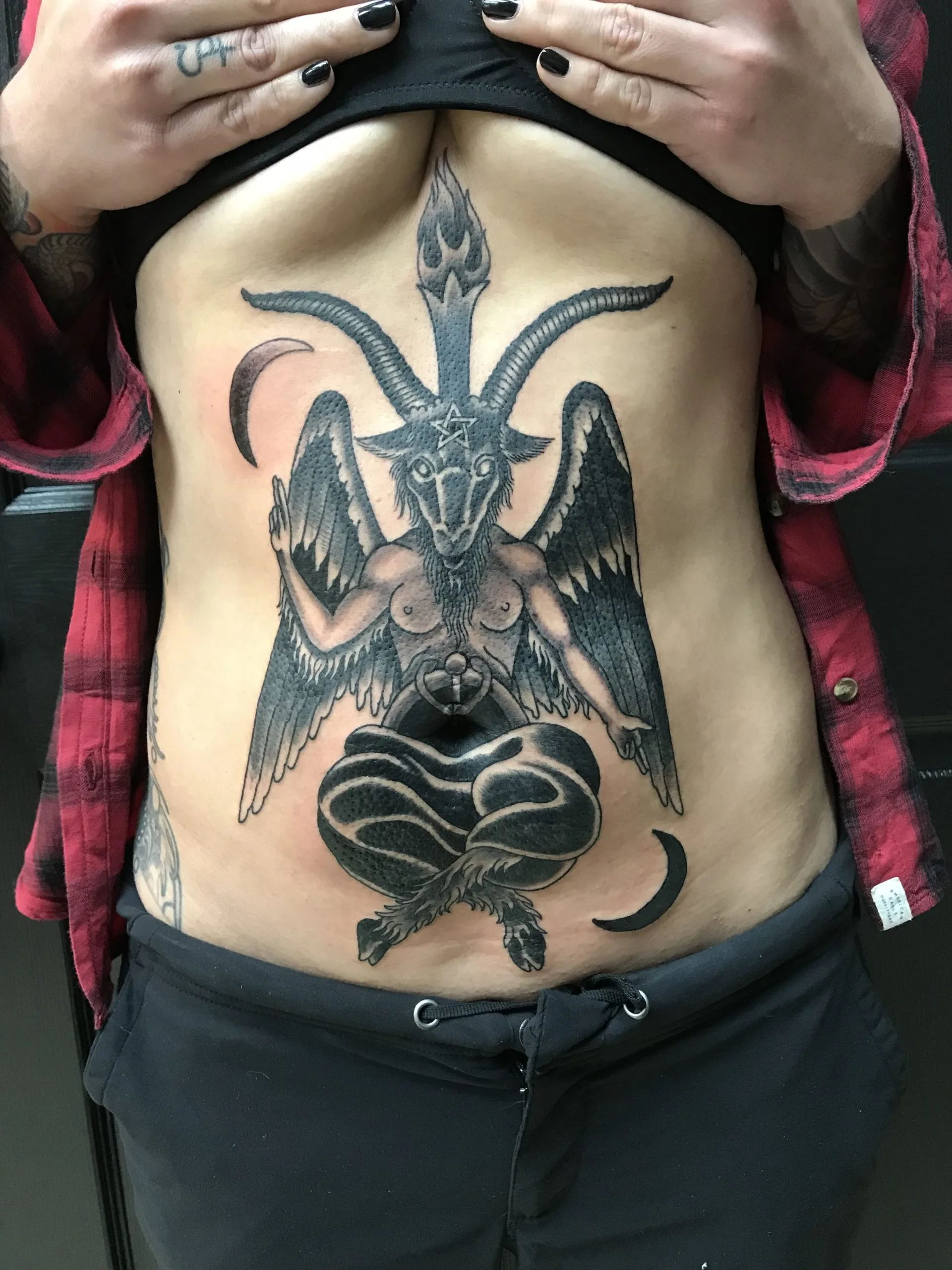 Winged goat-headed figure with horns over a heart shape, black and grey, on the abdomen with crescent moons on each side