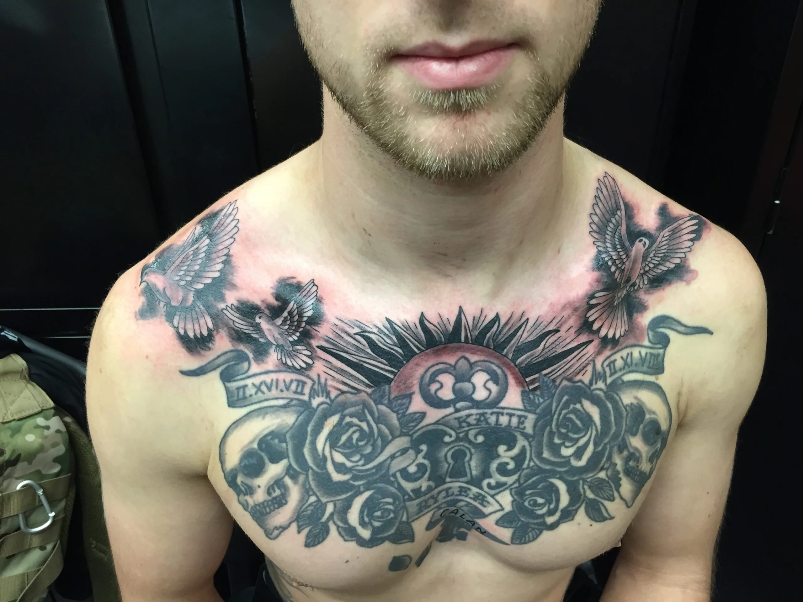 Chest piece with a skull, roses, a heart-shaped lock banner, birds, and sunburst rays in black and grey