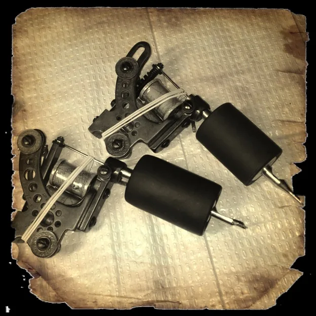 Pair of tattoo machines with black frames and coils on a textured cloth, sepia-toned photo