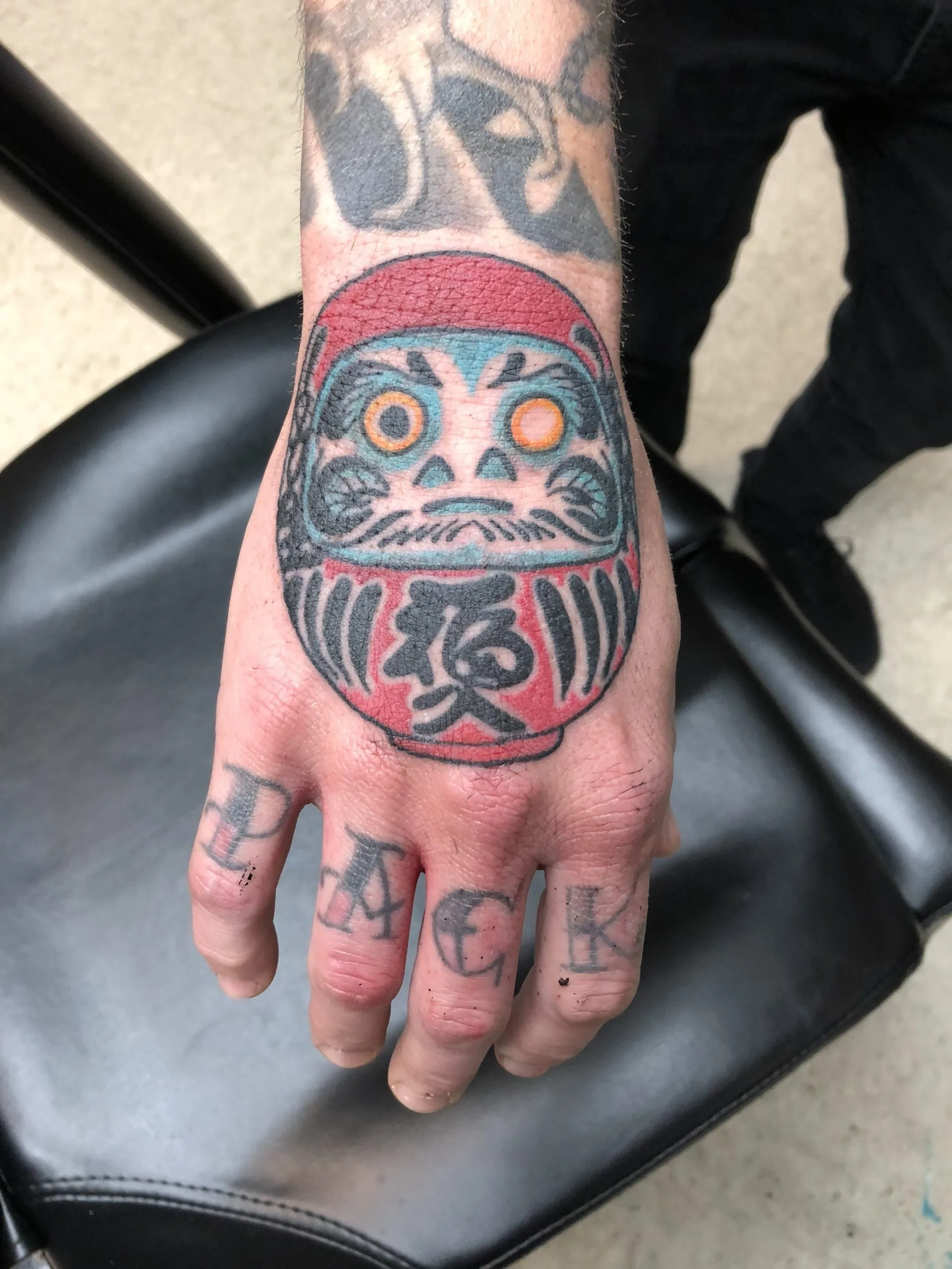Daruma doll tattooed on the back of a hand in red, teal and black, with kanji on the body and bold lettering across the knuckles