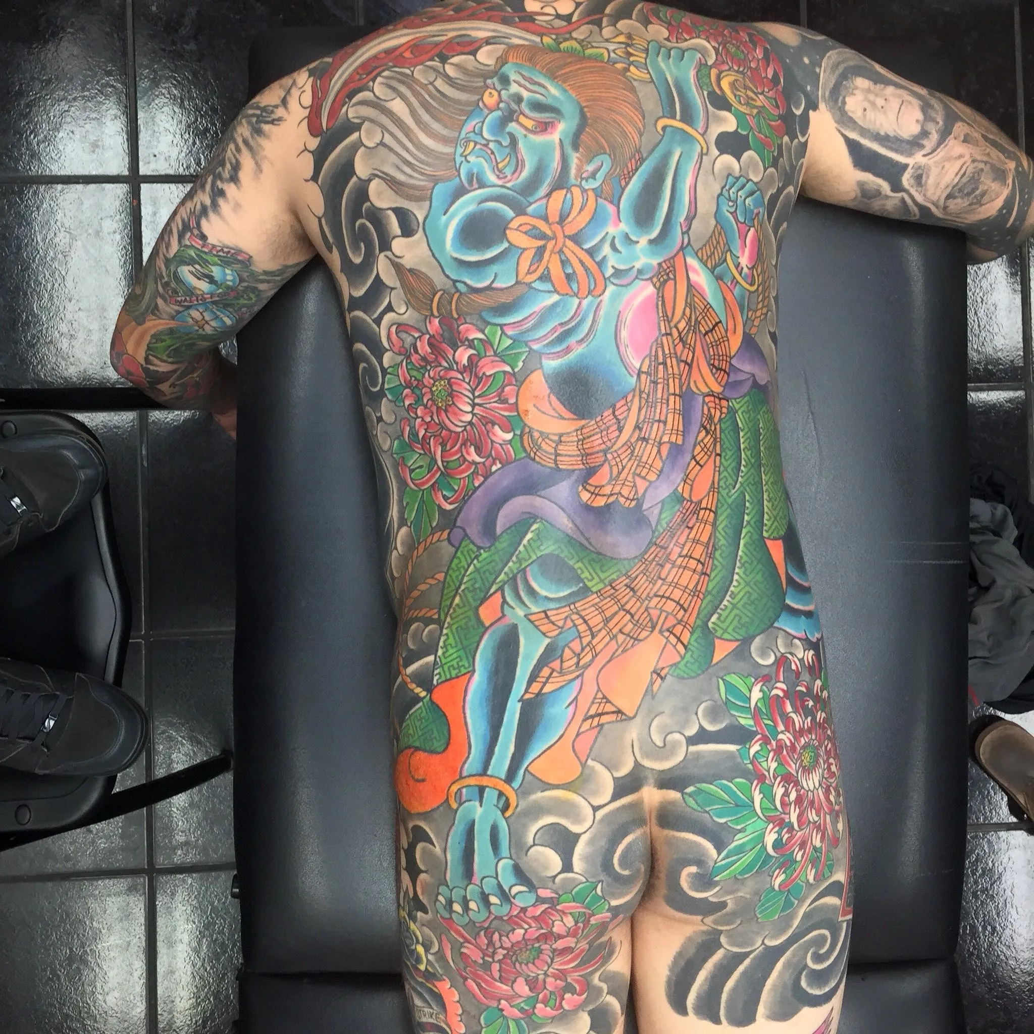 Full back piece of a large blue-skinned deity figure in flowing green and purple robes, framed by red chrysanthemums and grey clouds