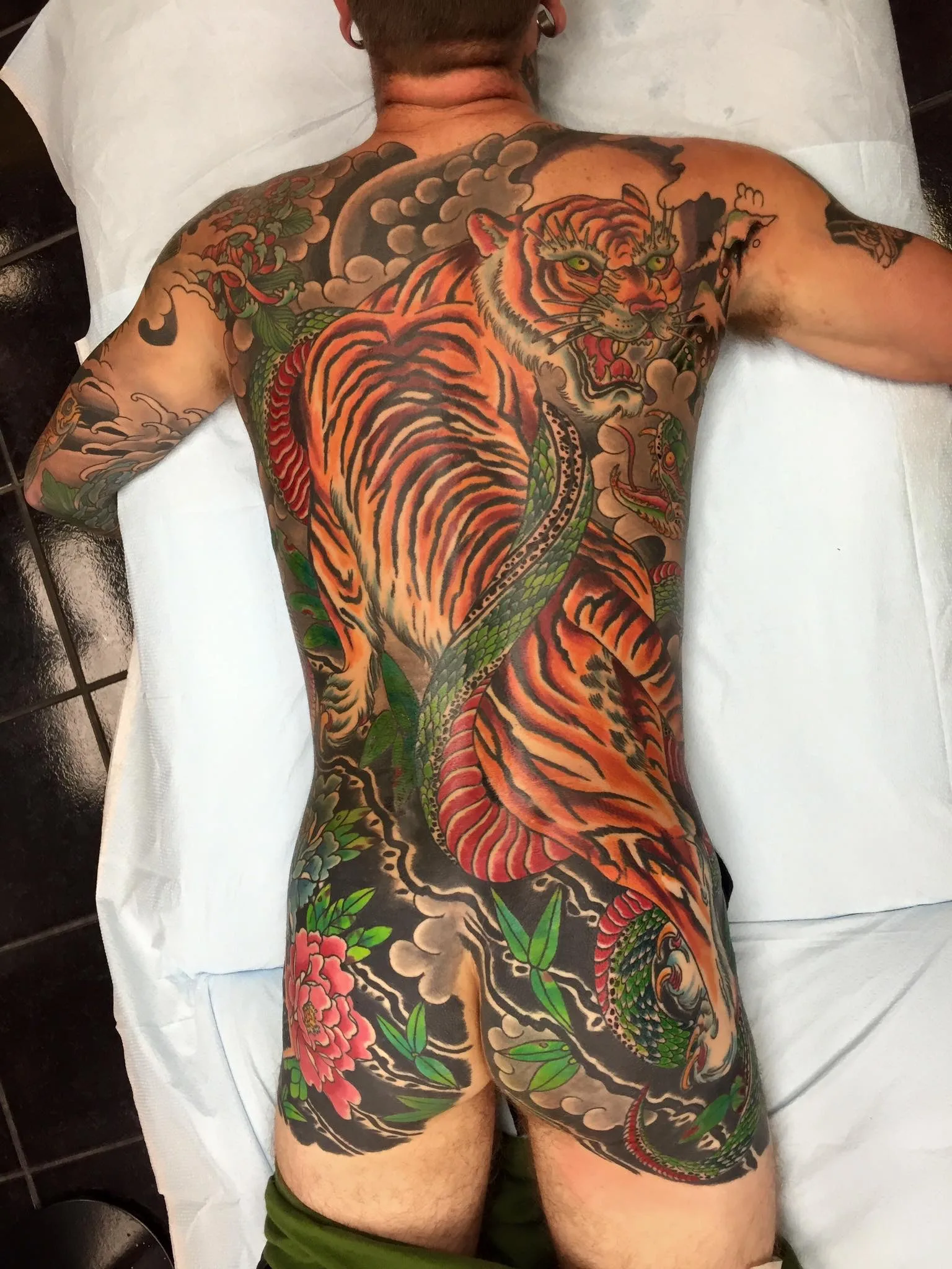 Full back piece of an orange and black tiger coiled with a green snake, over a red peony and grey clouds
