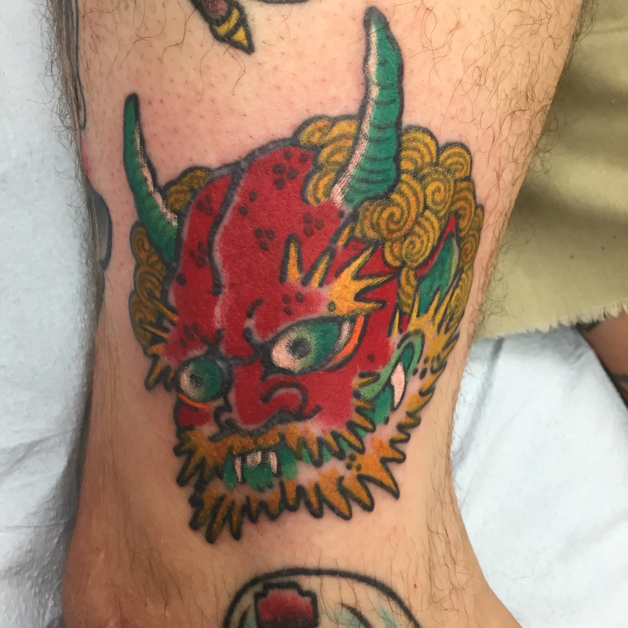 Red horned mask with green horns and yellow flame-like hair, tattooed on a lower leg