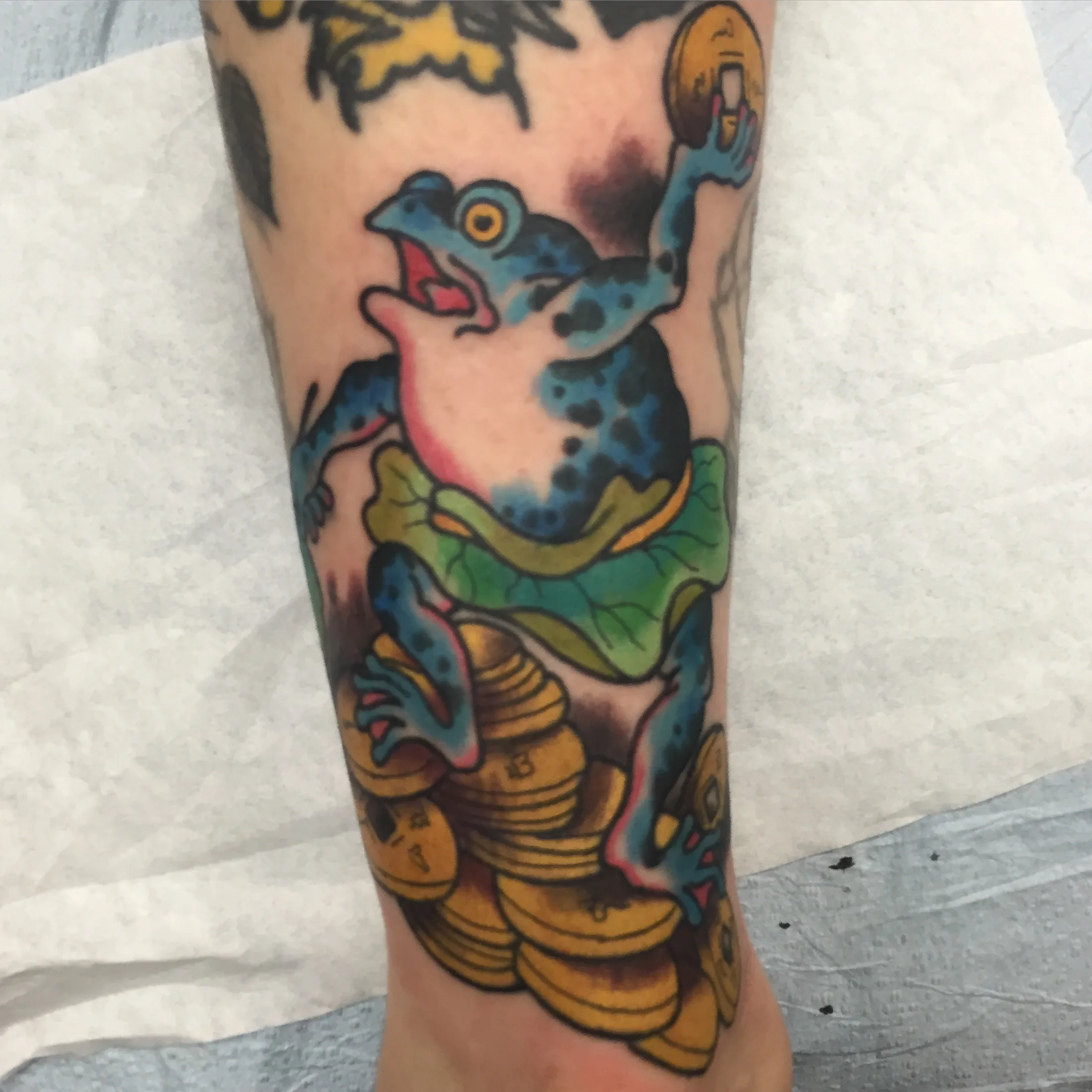 Blue frog perched on a green lotus pad above a stack of gold coins, tattooed on a lower leg