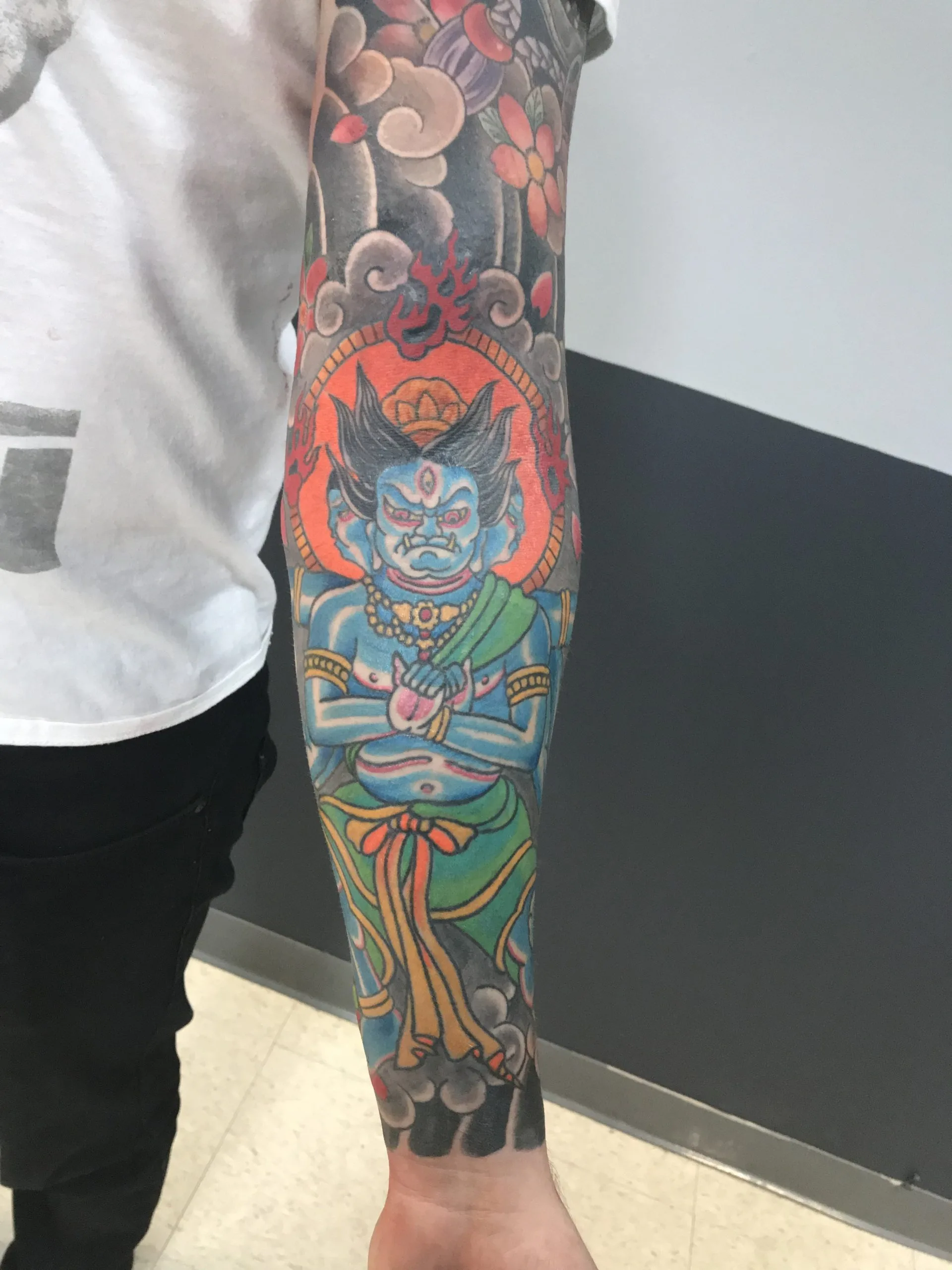 Colour Japanese sleeve of a blue-skinned deity seated before an orange halo, surrounded by red blossoms and grey clouds