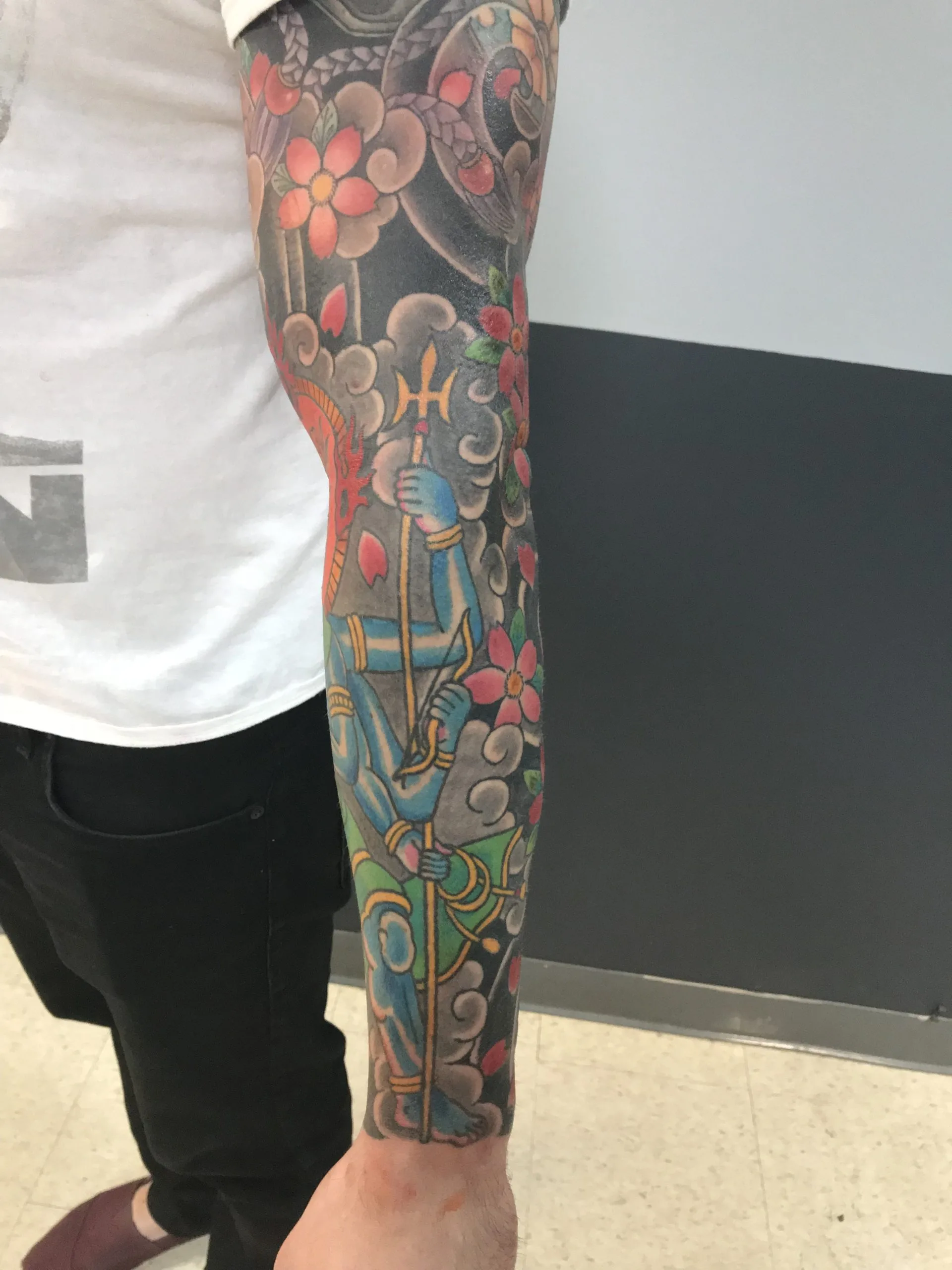 Colour Japanese sleeve with a blue-skinned figure holding a long staff on the forearm, among pink blossoms and grey clouds