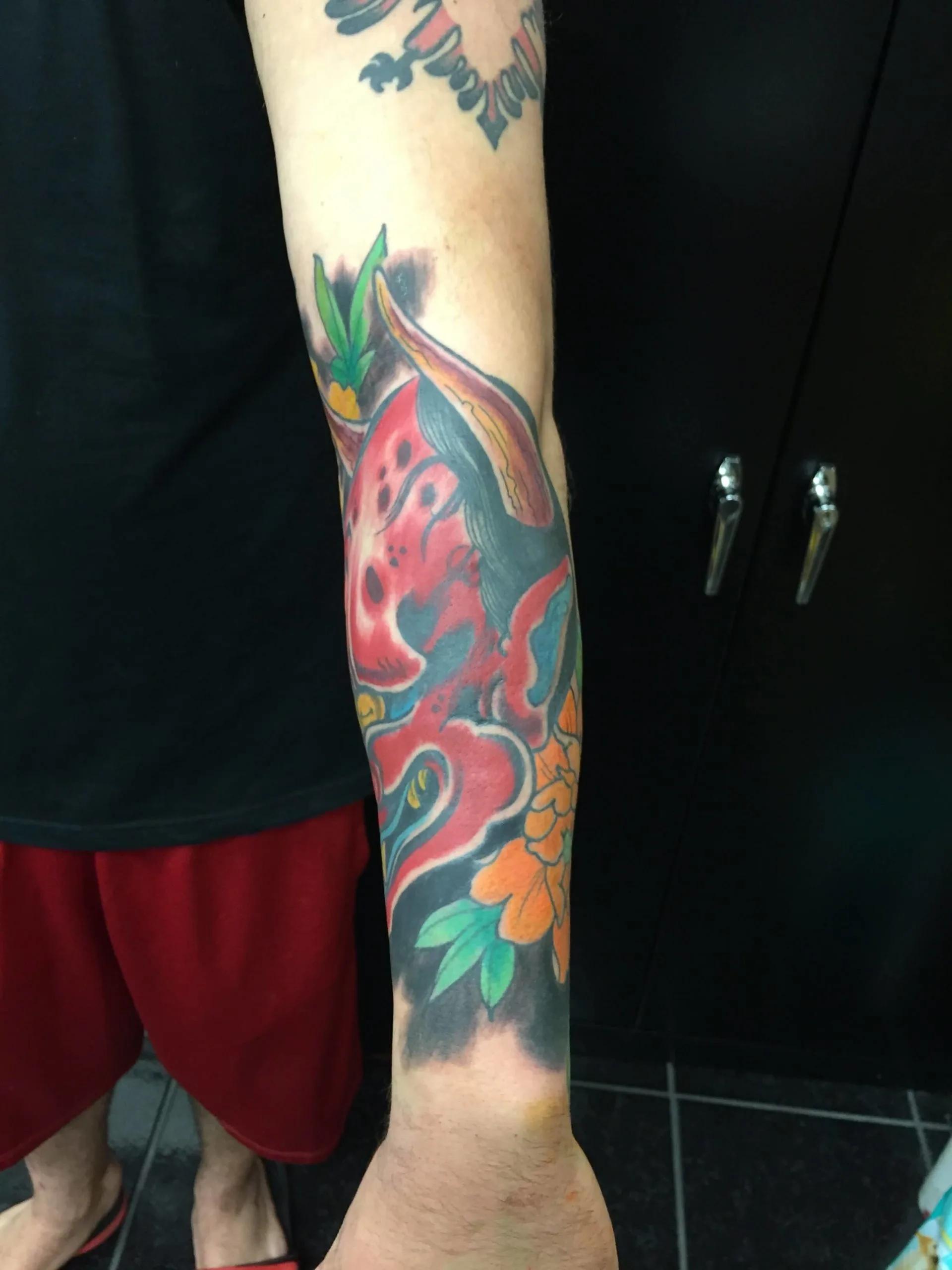 Forearm tattoo with a stylized red and teal face-like design surrounded by orange flowers and green leaves, color