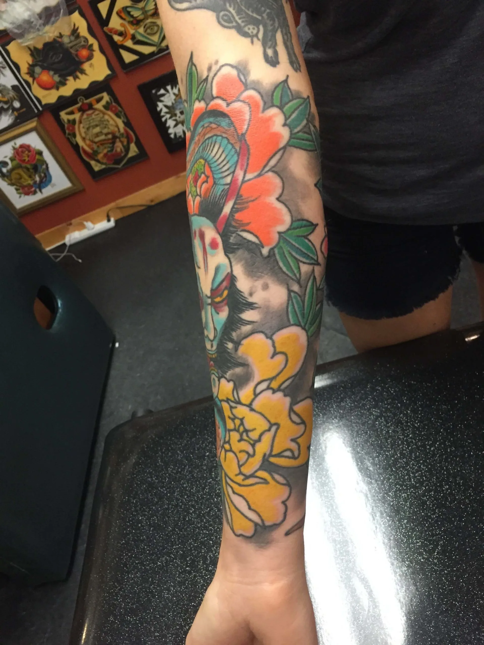 Color forearm tattoo of a teal mask-like face between orange and yellow peonies over black and grey shading