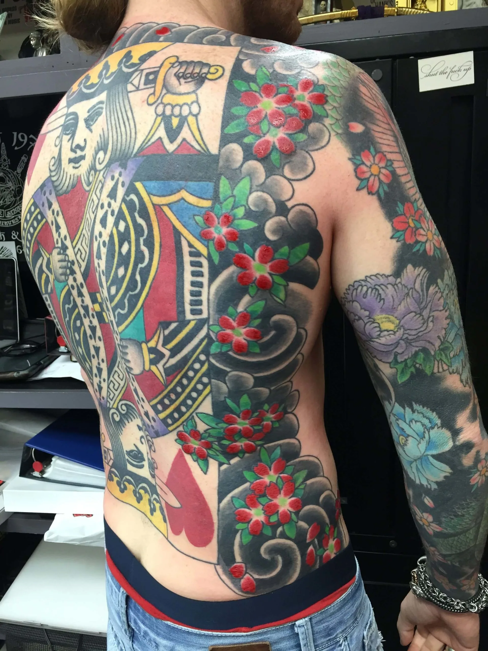 Full back piece with a king playing-card figure and grey cloud scrollwork with red blossoms, extending into a peony arm sleeve
