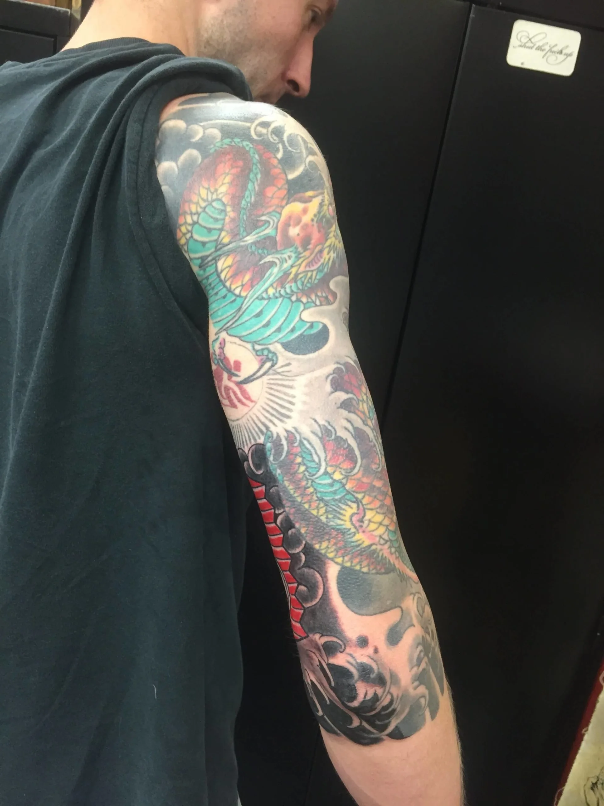 Teal and red Japanese dragon winding down a full arm sleeve amid grey clouds and waves