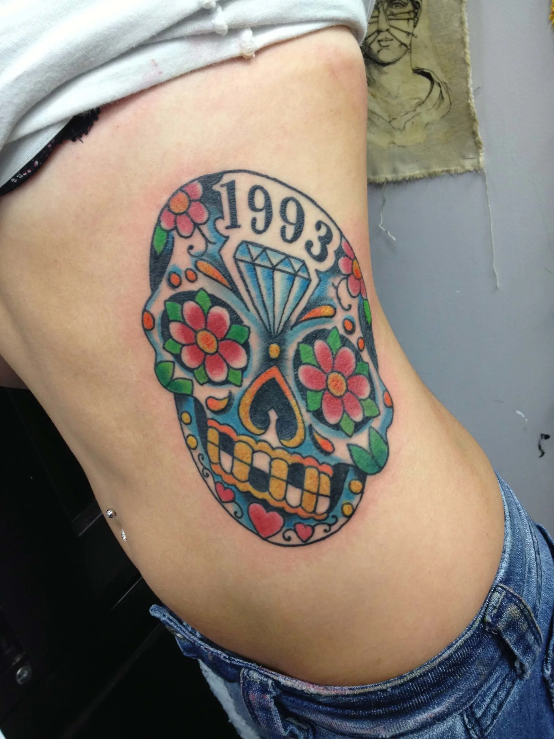Ribs tattoo of a colorful sugar skull with a diamond, flowers, and '1993' across the forehead