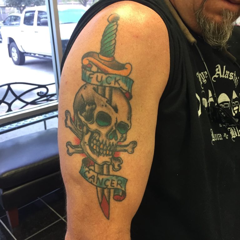 Color upper arm tattoo of a skull and crossbones pierced by a dagger, with a banner reading Fuck Cancer