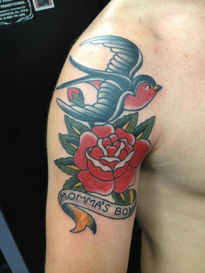 Upper arm tattoo of a swallow in flight above a red rose and a 'MOMMA'S BOY' banner