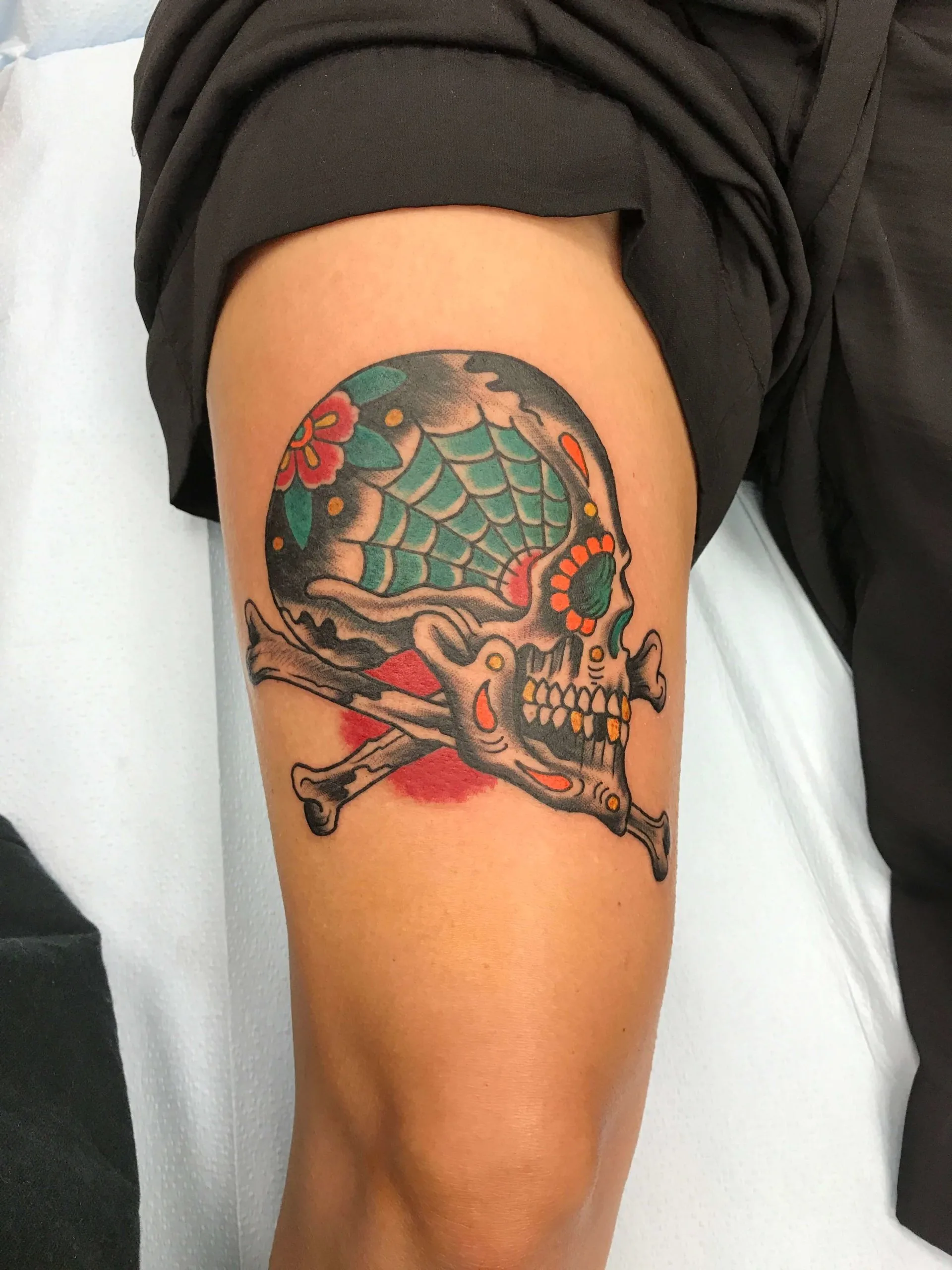 Skull and crossbones thigh tattoo with a teal spiderweb pattern and orange flowers