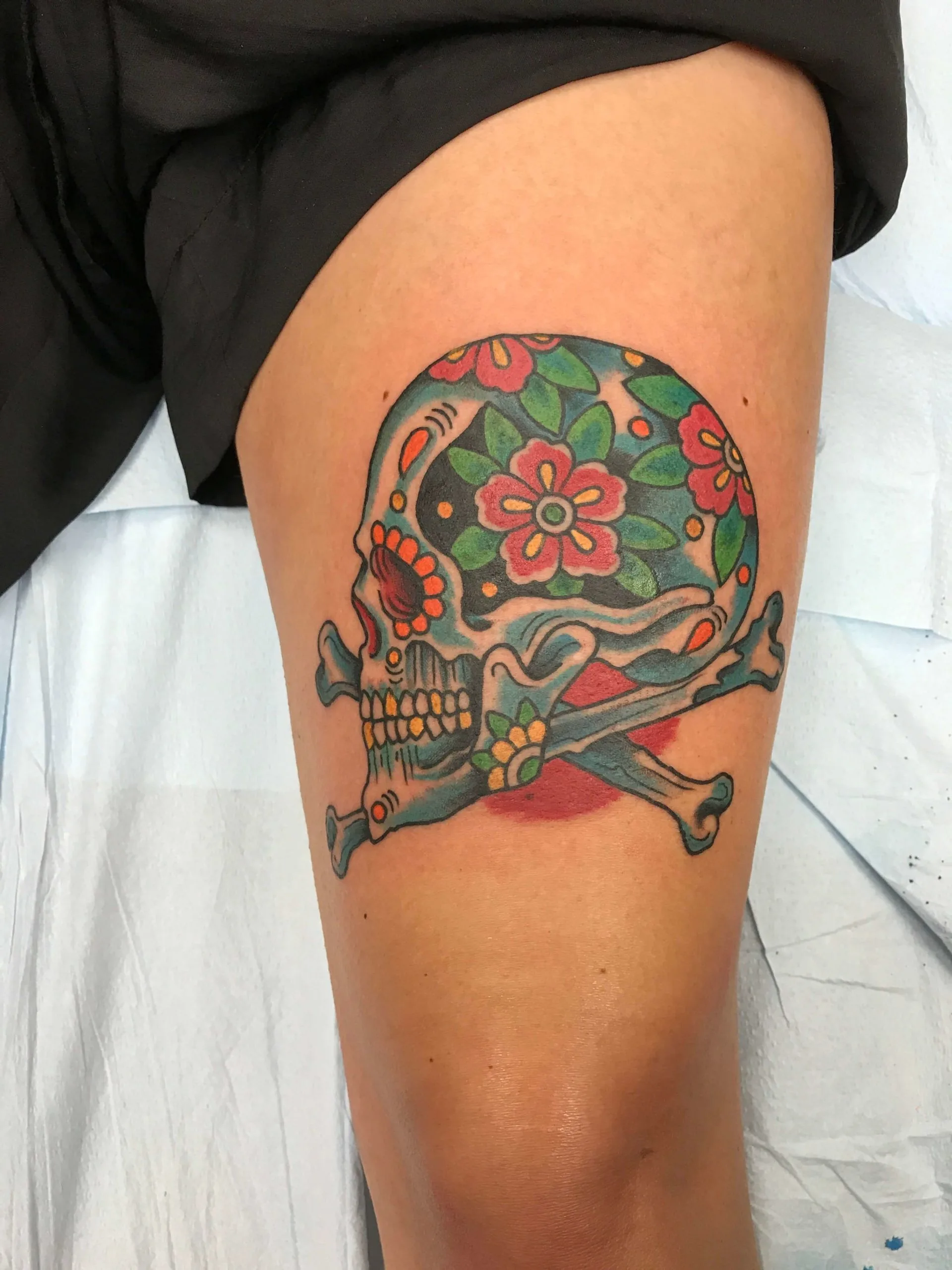 Colour thigh tattoo of a sugar-skull-style skull and crossbones with pink and red flowers