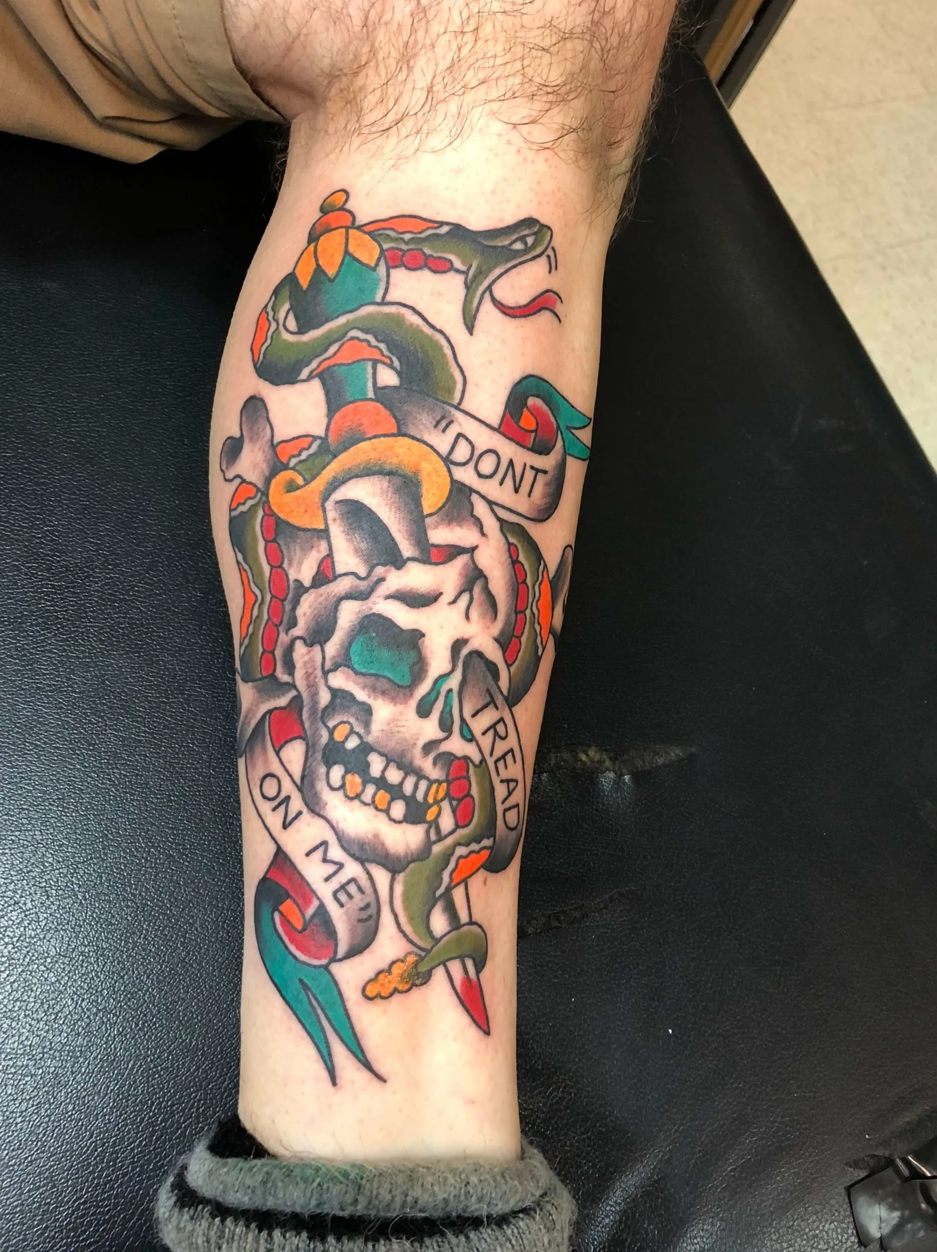 Lower leg tattoo of a grey skull pierced by a dagger, wrapped by a green snake, with a Don't Tread on Me banner