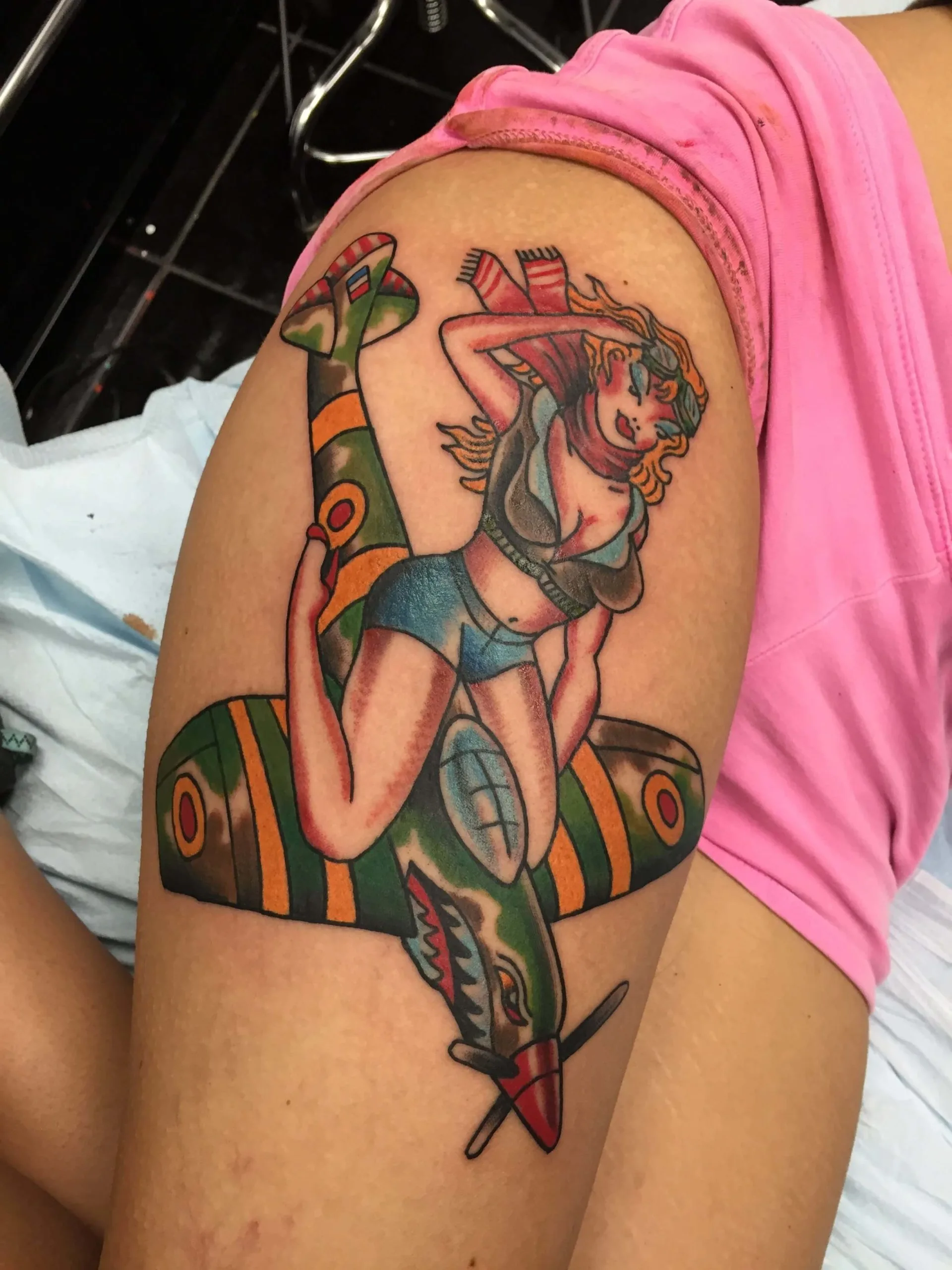 Color upper arm tattoo of a pin-up woman in a blue outfit and red headscarf posed on a green and orange fighter plane
