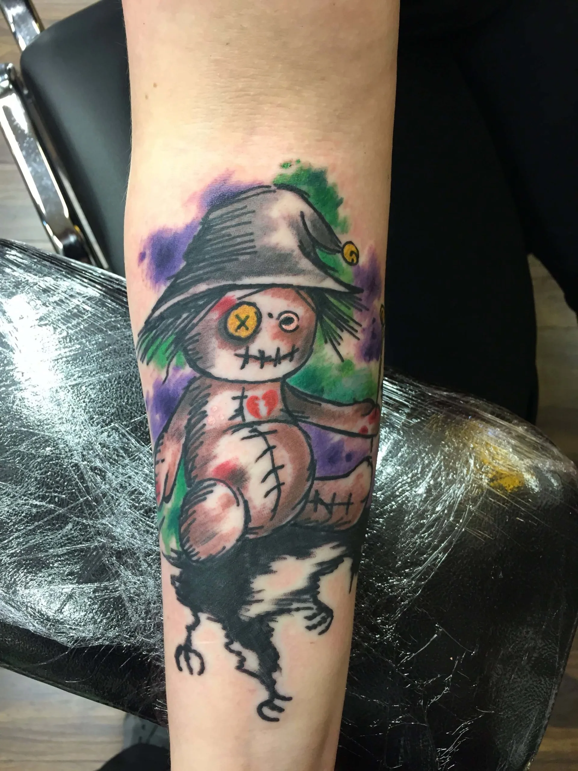 Colour forearm tattoo of a stitched scarecrow-like doll on a purple and green watercolor wash background