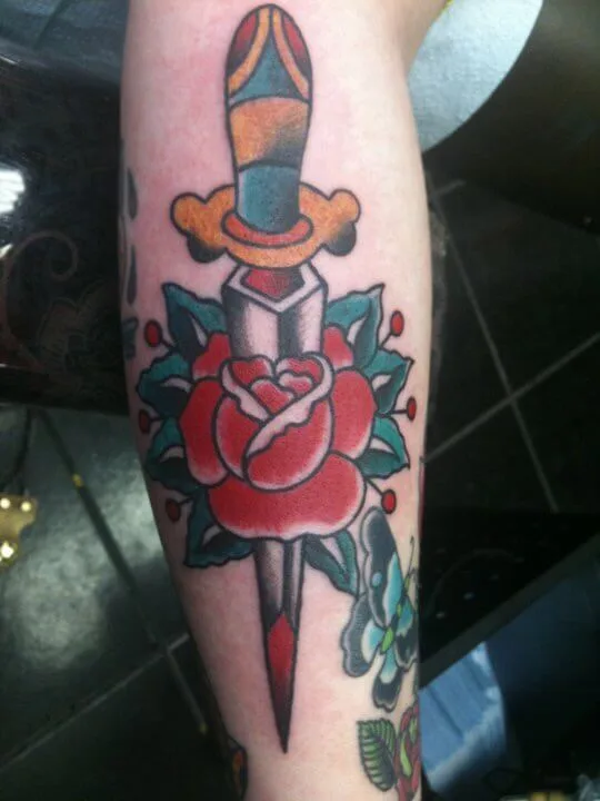 Ornate dagger piercing a red rose with green leaves and a small blue butterfly, forearm