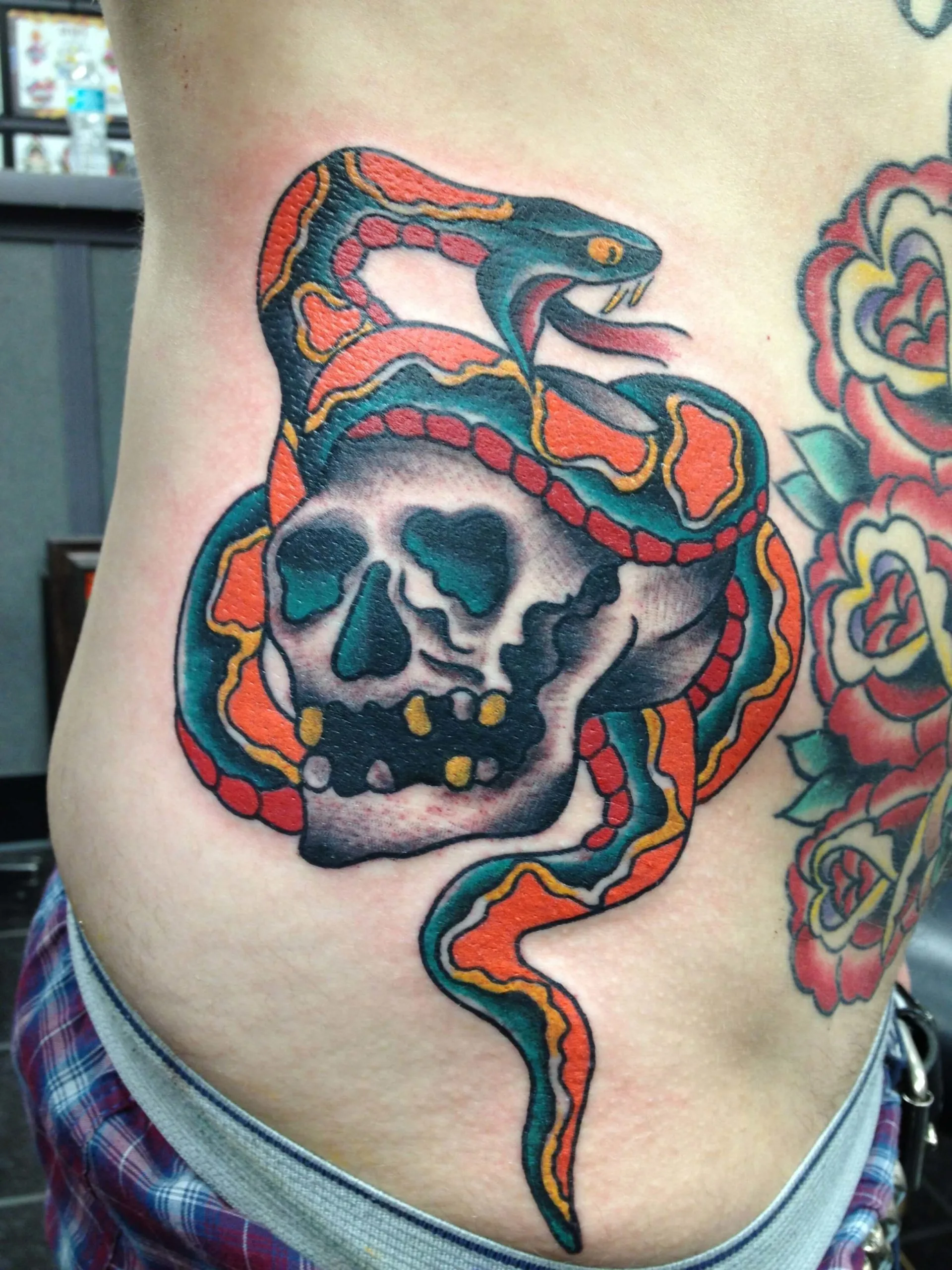 Colour ribs tattoo of a snake coiled around a skull with red roses beside it