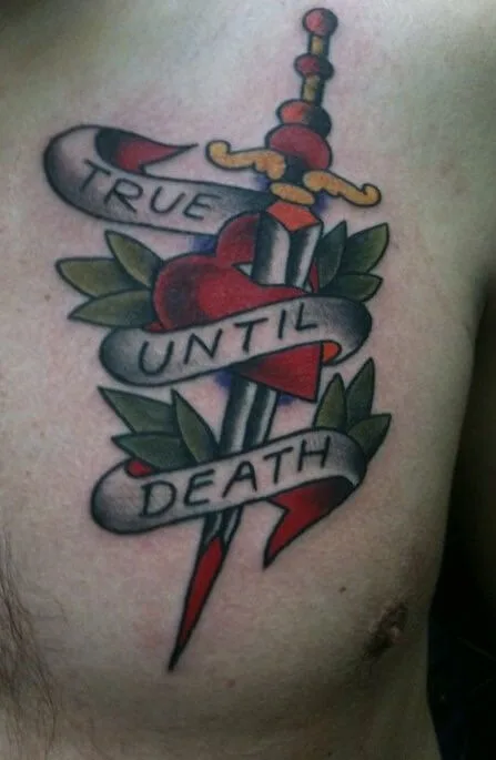 Color tattoo of a dagger through a red heart with banners reading 'TRUE UNTIL DEATH' and green leaves, on the ribs