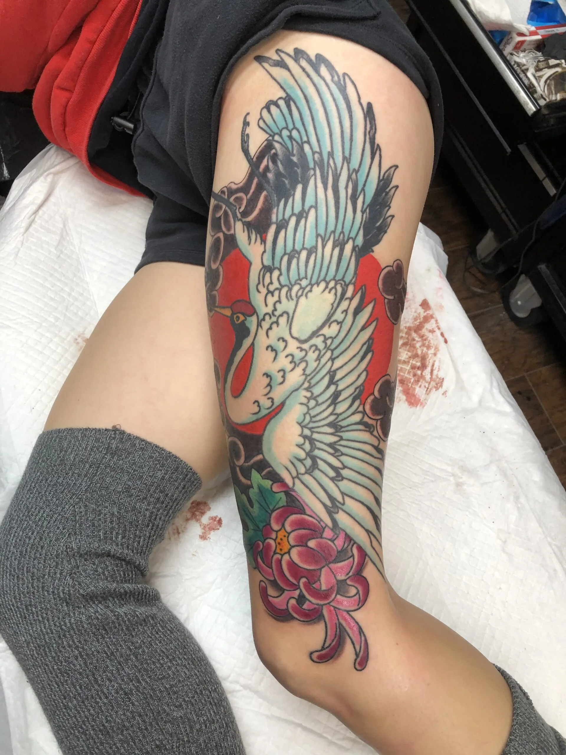 Colour leg tattoo with a white and teal crane in flight, red clouds, pink peony near the ankle
