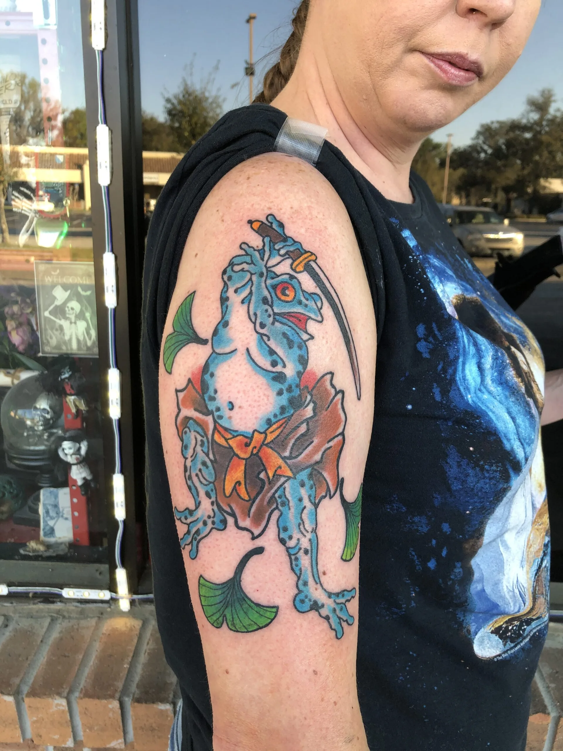 Colour upper arm tattoo of a blue frog character wielding a katana with an orange sash and ginkgo leaves