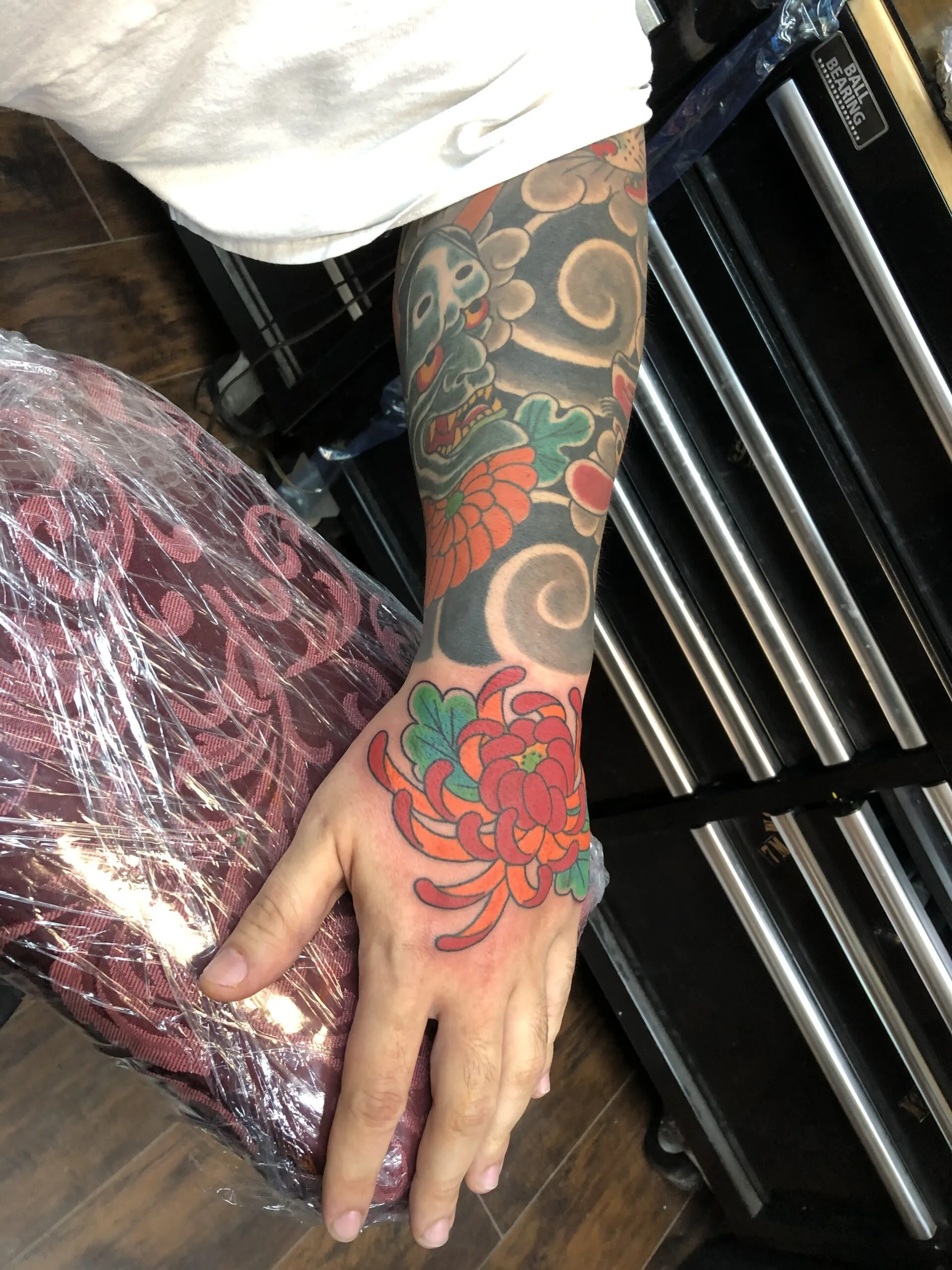Grey demon mask with orange accents on the forearm above a red chrysanthemum on the hand