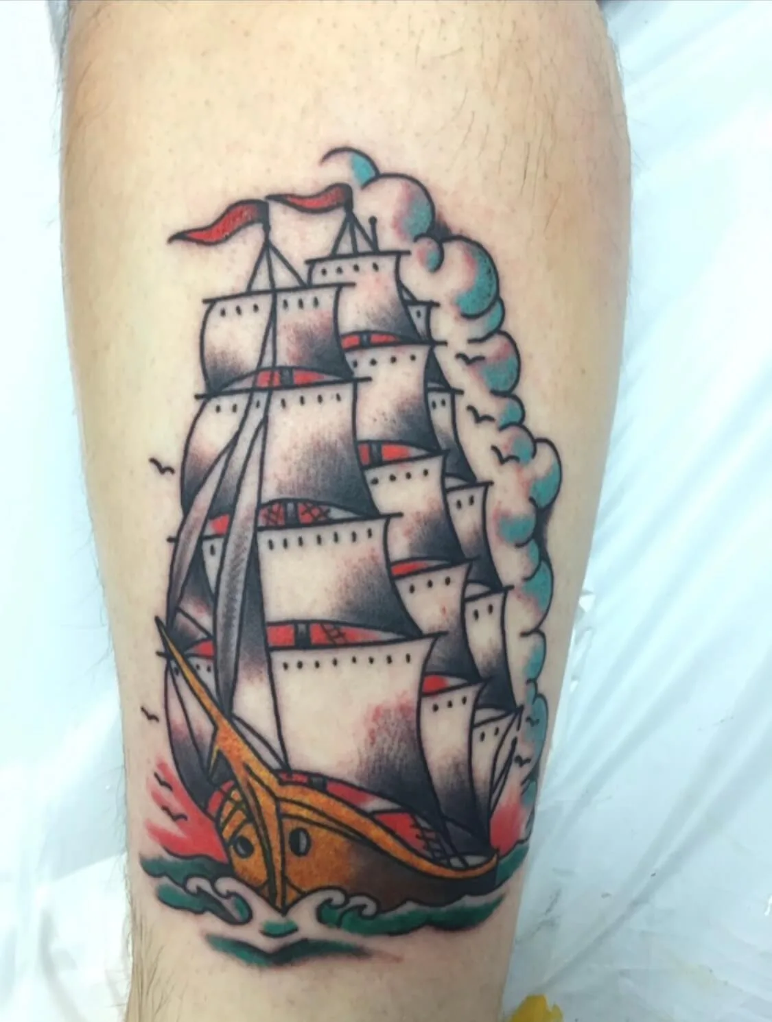 Calf tattoo of a black-and-red tall sailing ship with teal smoke clouds and wave details below