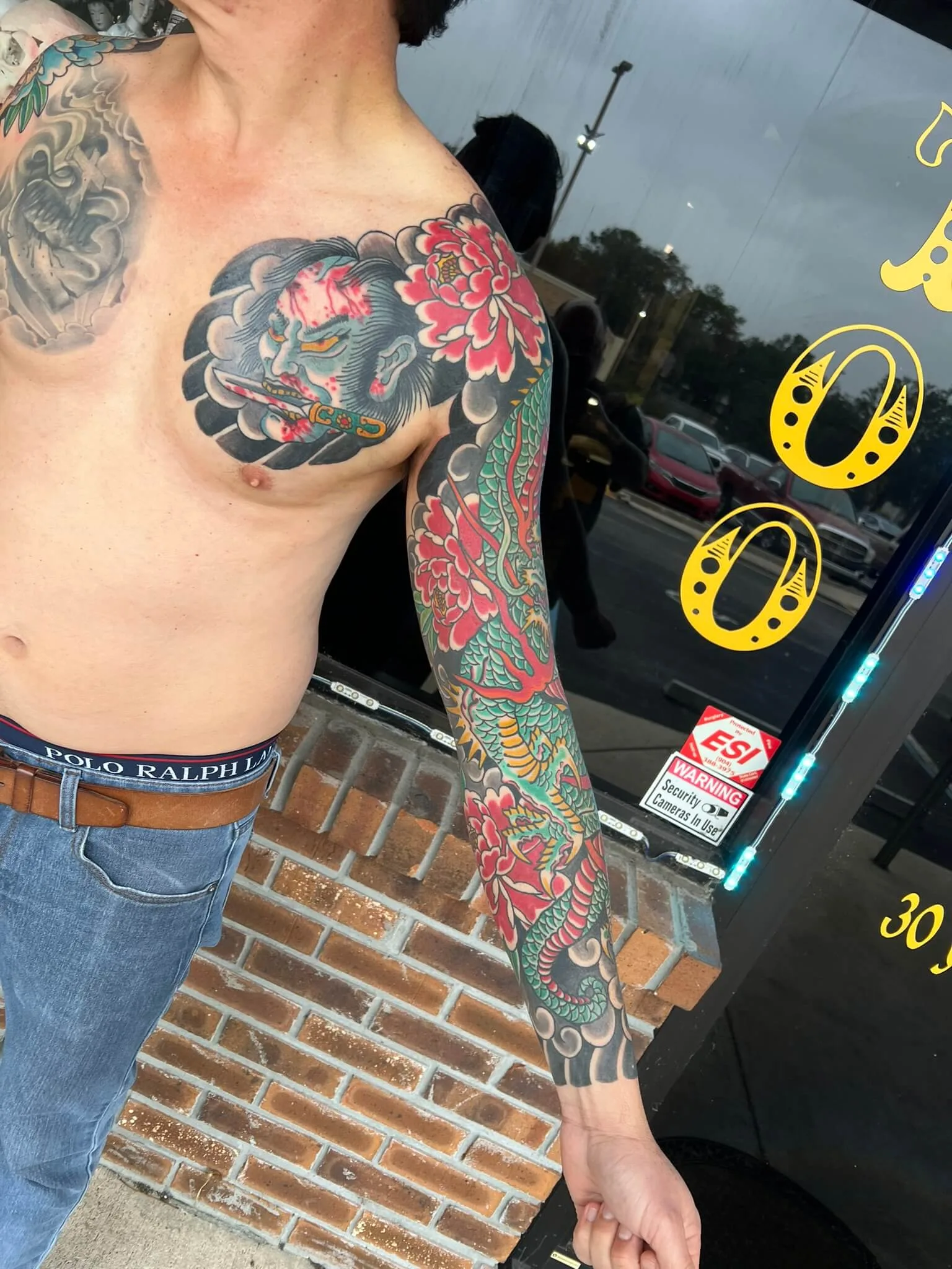 Chest panel of a blue-grey demon mask biting a dagger beside a red peony, connecting to a green dragon sleeve down the arm