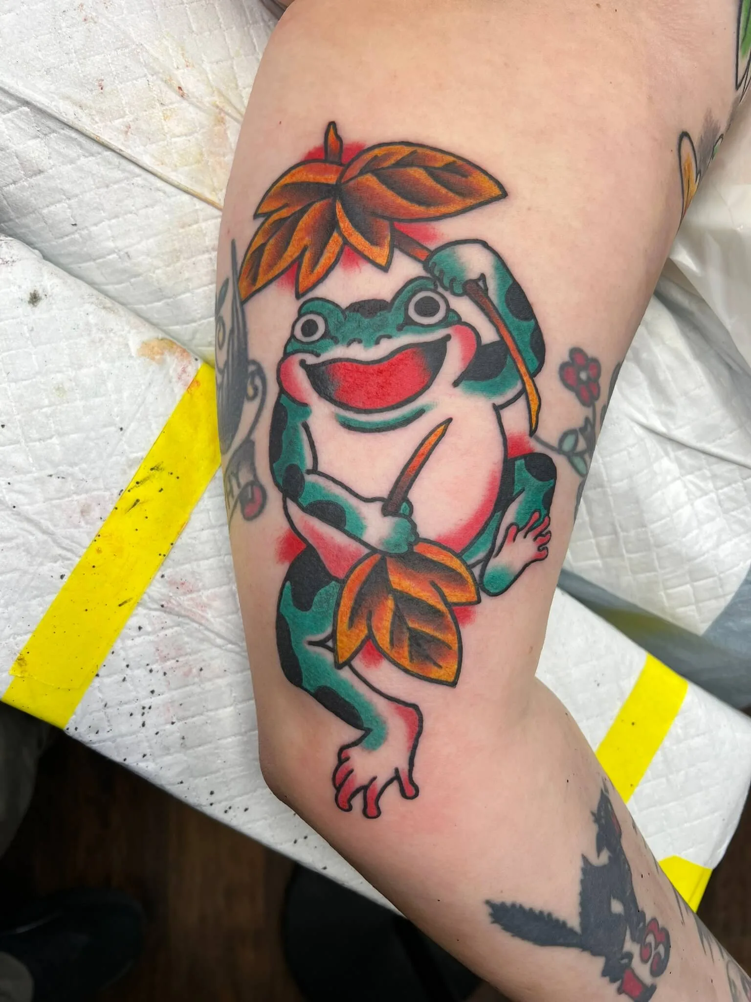 Inner upper arm tattoo of a green cartoon frog holding a leaf umbrella and a knife, with maple leaves