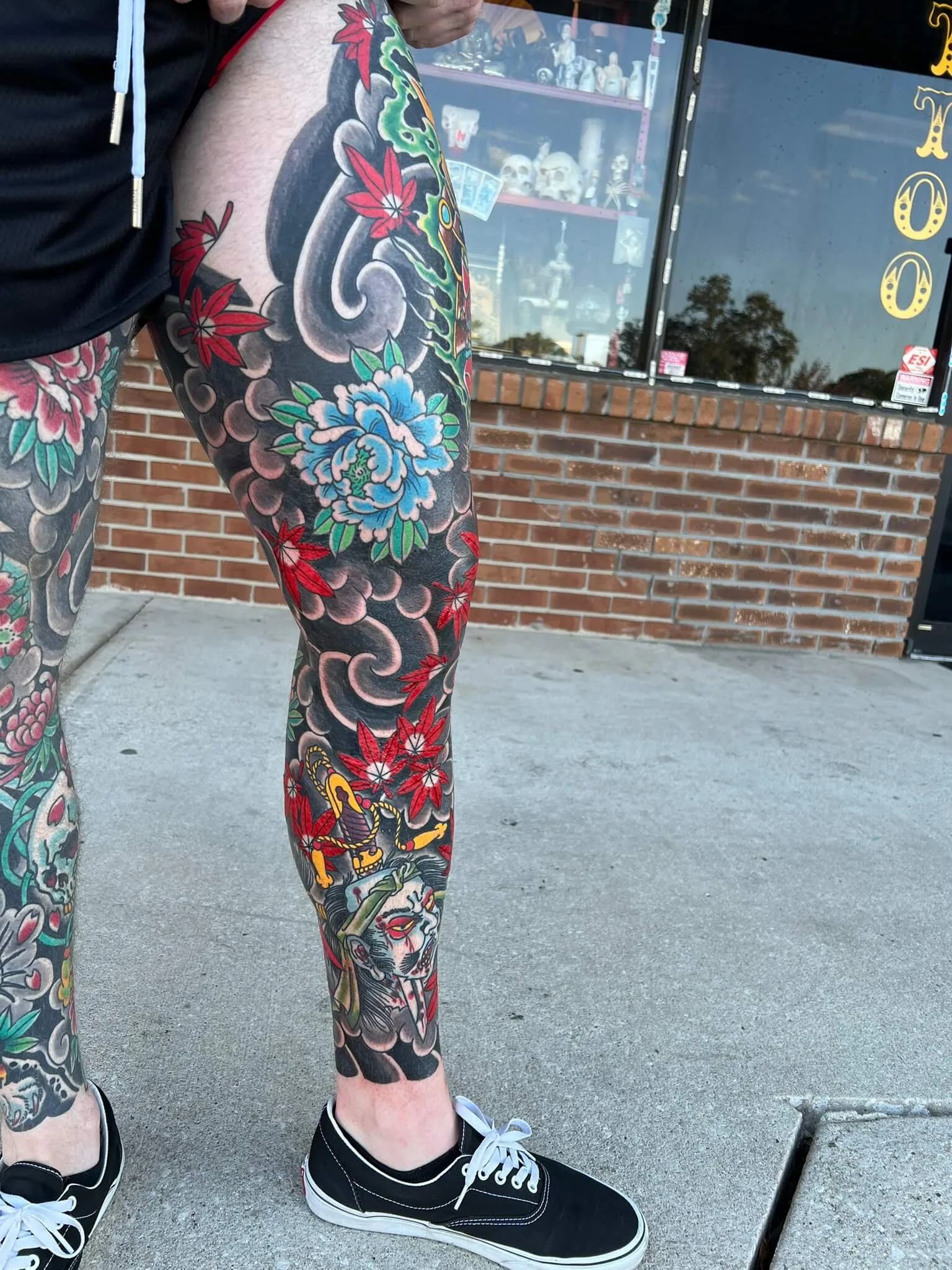 Full leg sleeve of grey cloud-and-wave work with red maple leaves, a blue peony, and a face gripping a dagger near the ankle