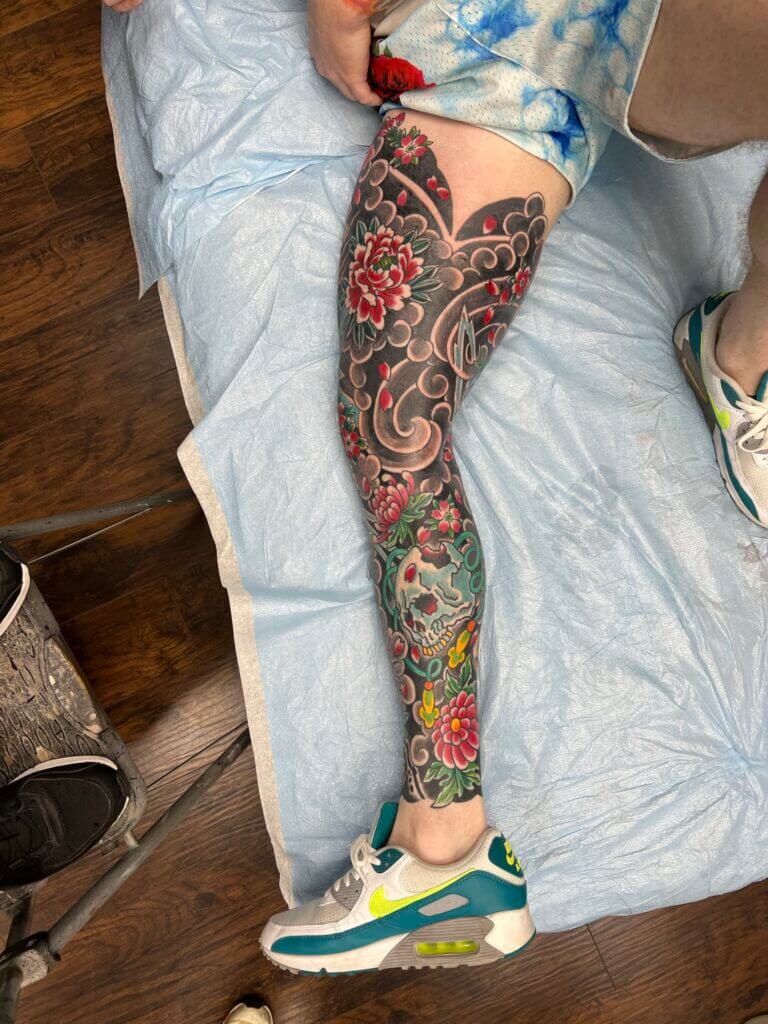 Colour lower leg tattoo with pink peonies, grey wave scrollwork, and a teal skull among green leaves